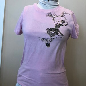 May include: A light pink t-shirt with a black and gold graphic of a robot with a round head and many gears.