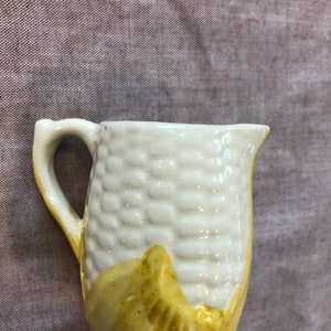 May include: A small white ceramic creamer jug shaped like an ear of corn with yellow painted details.