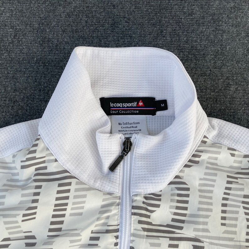 May include: White golf jacket with a zipper closure and a black le coq sportif logo on the front. The jacket has a geometric pattern in gray and white.