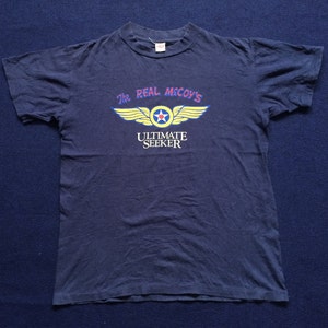 May include: A navy blue t-shirt with a yellow and red graphic of a winged star with the text "The Real McCoy's Ultimate Seeker".