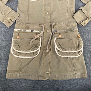 May include: A green utility jacket with a drawstring waist and two large pockets with white trim. The jacket has a button closure and a camouflage pattern on the pockets.