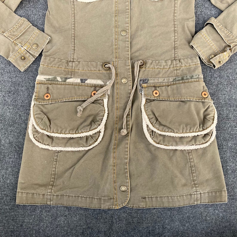 May include: A green utility jacket with a drawstring waist and two large pockets with white trim. The jacket has a button closure and a camouflage pattern on the pockets.