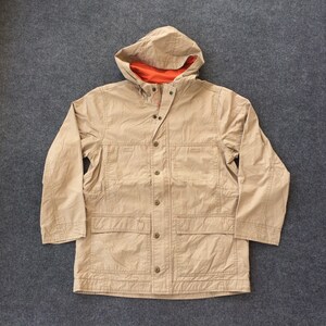 Puede incluir: Una chaqueta beige con capucha, cierre de botones y varios bolsillos. La capucha tiene un forro naranja contrastante.