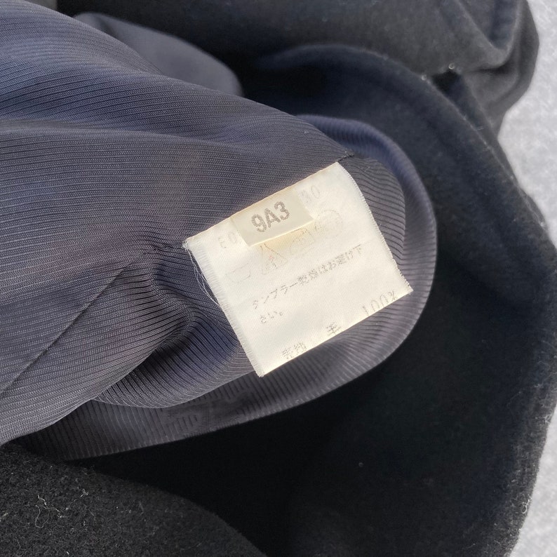 May include: Close-up of a black garment's interior, revealing a dark blue lining. A white tag displays text and symbols, including "9A3" and "100%" in Japanese. The garment's fabric appears to be wool.
