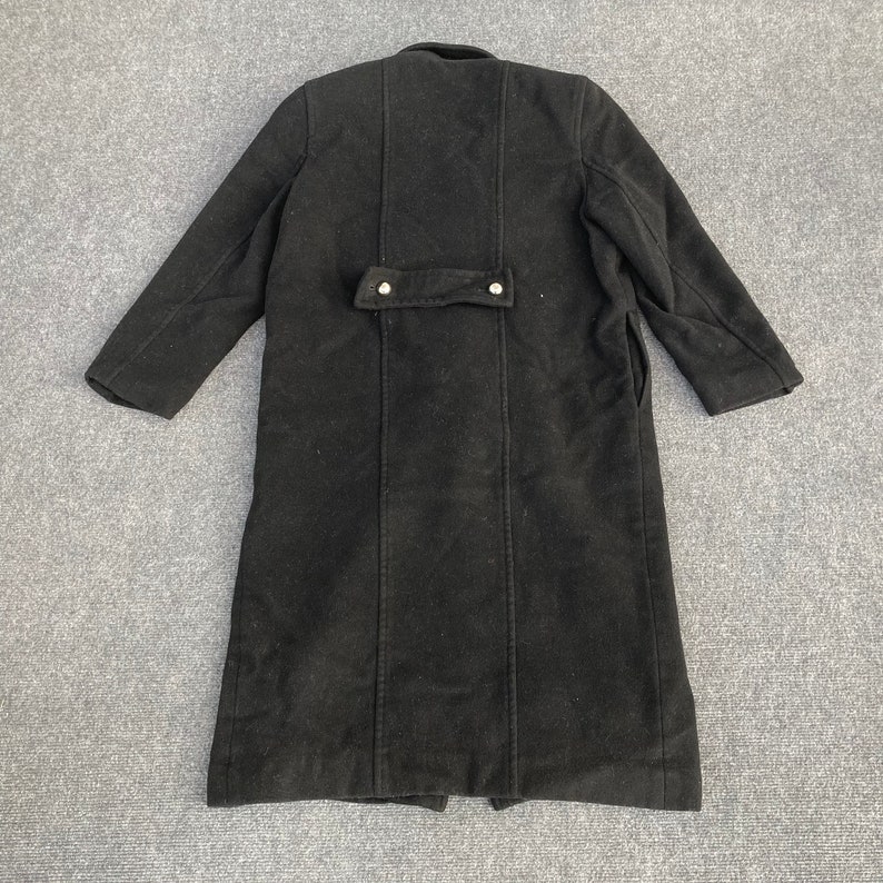 May include: A long, black coat with a buttoned back detail and a notched collar. The coat has long sleeves and a tailored fit, suitable for colder weather. The coat is made of a thick, dark fabric.
