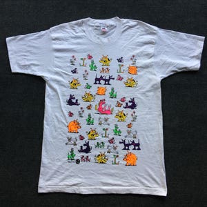 May include: White t-shirt with a colorful all-over print of cartoon dogs in various poses. The dogs are in shades of pink, orange, yellow, green, purple, and black.