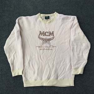 Puede incluir: Una sudadera de color rosa claro con mangas largas y cuello y puños de canalé de color crema. La sudadera tiene un logotipo MCM bordado en marrón con el texto "mode creation munchen".