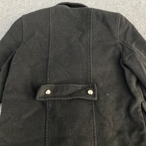 May include: A black wool coat with a buttoned flap detail at the lower back. The coat has a classic design with a tailored fit, featuring a collar and seams that run vertically down the back. The buttons are silver-colored.