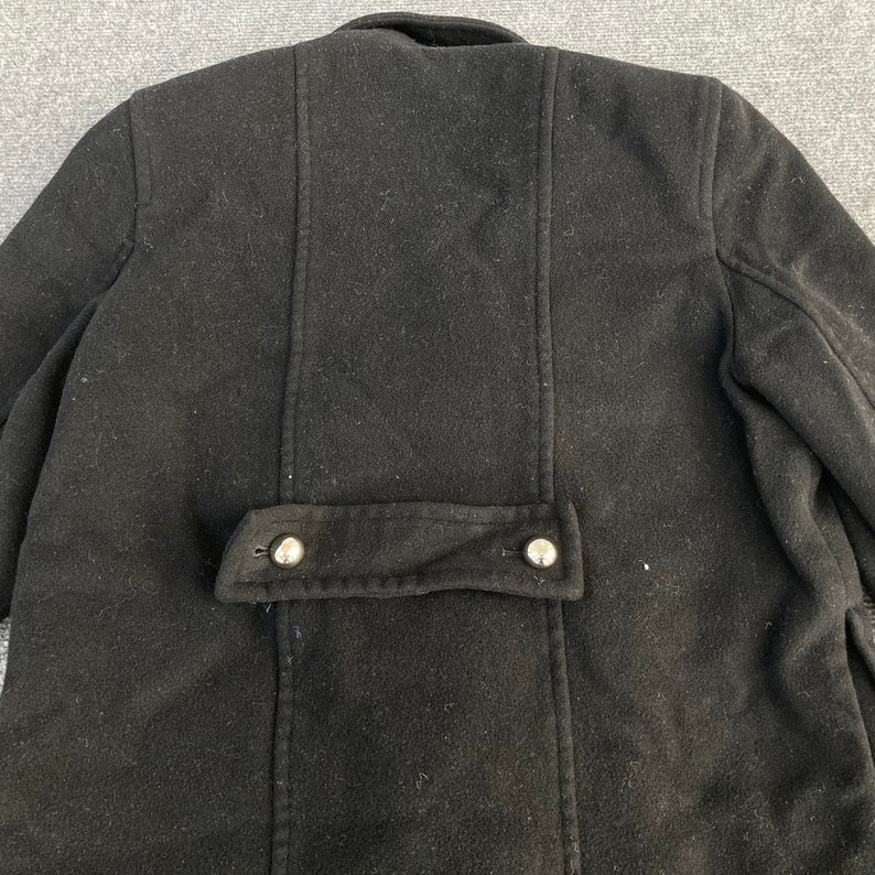 May include: A black wool coat with a buttoned flap detail at the lower back. The coat has a classic design with a tailored fit, featuring a collar and seams that run vertically down the back. The buttons are silver-colored.