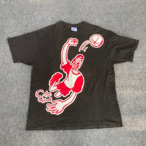 Puede incluir: Camiseta negra con una ilustración de dibujos animados roja y blanca de una persona jugando al baloncesto. La persona lleva una camiseta y pantalones cortos rojos y blancos. El balón de baloncesto es rojo y blanco.