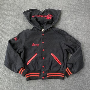 Puede incluir: Chaqueta de béisbol negra con ribetes rojos y una capucha bordada en rojo. La chaqueta tiene cierre de cremallera y un frente abotonado. La chaqueta tiene un nombre bordado en rojo "Stacey" en la parte delantera y el número "86" en la manga izquierda.