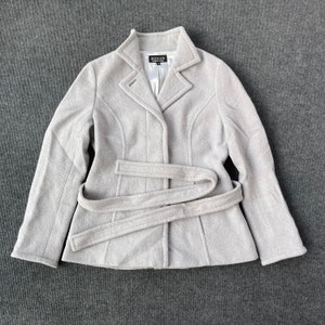 Può includere: Un cappotto di lana grigio chiaro con cintura e un'etichetta nera con la scritta "SONIA" sul davanti.