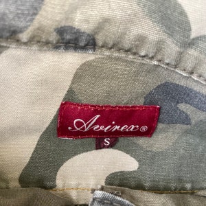 May include: A red label with the text "Avivex" in a cursive font and a size "S" printed below. The label is on a camouflage fabric background.