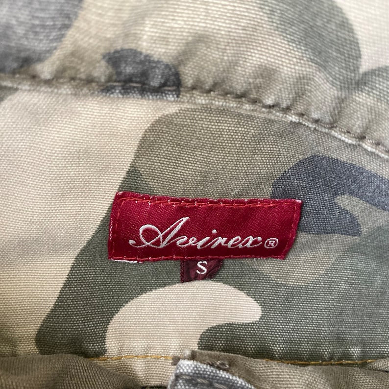 May include: A red label with the text "Avivex" in a cursive font and a size "S" printed below. The label is on a camouflage fabric background.