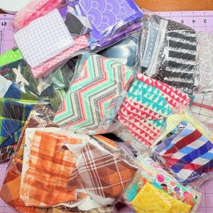 May include: A collection of fabric bundles in various colors and patterns, including plaid, chevron, and geometric designs. The fabrics are packaged in clear plastic, showcasing the assortment of textures and hues. The color palette includes pink, green, orange, and blue.