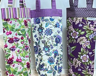 May include: Three fabric wine bottle bags with handles. The bags feature floral and patterned designs in purple, green, and white. One bag has a fuchsia flower print, another has blue floral, and the third has purple floral. Each bag has a coordinating handle.
