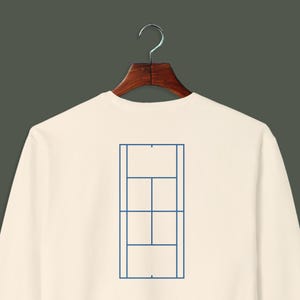 May include: Cream-colored sweatshirt with a blue tennis court diagram on the back. The diagram features the court's lines and dimensions, creating a minimalist design. The sweatshirt is hanging on a wooden hanger.