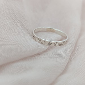 May include: A silver ring with etched celestial designs, including stars, moons, and planets. The ring is set against a soft, pink fabric background. The ring's band is approximately 0.2 cm wide.