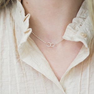 May include: A silver chain necklace with two interlocking circles as a pendant. The necklace is worn by a person with a light beige shirt.