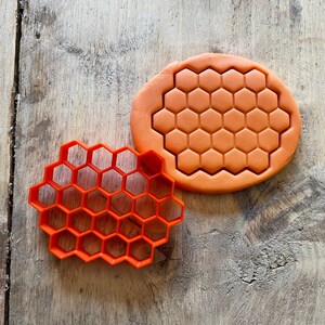 May include: Two orange plastic cookie cutters shaped like honeycombs. The larger cutter is solid and the smaller cutter is hollow.