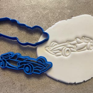 Puede incluir: Un cortador de galletas de pl&aacute;stico azul con forma de coche de carreras se encuentra junto a un trozo de masa blanca con una huella de coche. El cortador es un contorno detallado de un coche de carreras.