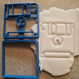 Op de afbeelding: Een blauwe plastic koekjesvormer in de vorm van een floppy disk. De vormer bestaat uit twee delen, een voor de omtrek en een voor de binnendetails. Een witte fondantkoek wordt met de vormer uitgesneden.