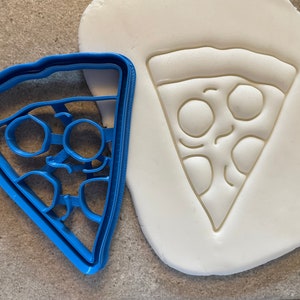 May include: A blue plastic cookie cutter shaped like a slice of pizza with pepperoni and cheese. The cutter is shown next to a white fondant impression of the pizza slice.