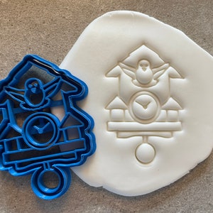 May include: Blue plastic cookie cutter shaped like a cuckoo clock with a bird on top. The cutter is next to a white fondant imprint of the same design.
