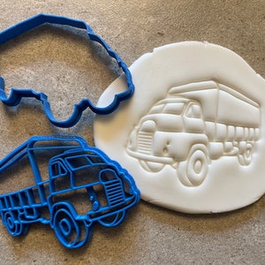 May include: Two blue plastic cookie cutters in the shape of a delivery truck. One cutter is a flat outline of the truck, and the other is a more detailed design with a raised bed and wheels. The cutters are on a white surface.