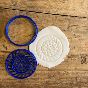 Könnte beinhalten: Zwei blaue Plastik-Ausstecher auf einer Holzoberfläche. Ein Ausstecher ist ein Kreis und der andere ist ein detailliertes Design mit einem Kreis in der Mitte und einem Wirbelmuster darum herum.