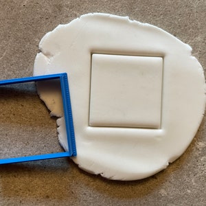 May include: A blue plastic square cookie cutter on a white fondant surface. The cutter has been pressed into the fondant, creating a square impression.