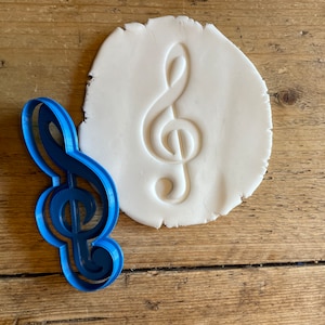 May include: A blue plastic cookie cutter shaped like a treble clef, used to cut out shapes from dough. The cutter is next to a white dough imprint of the treble clef.