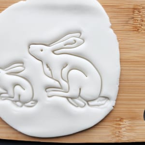 May include: A white, circular dough with two rabbit impressions, one large and one small, sits on a wooden cutting board. The rabbits are in profile, with detailed features. The board has a rounded corner and a handle hole.