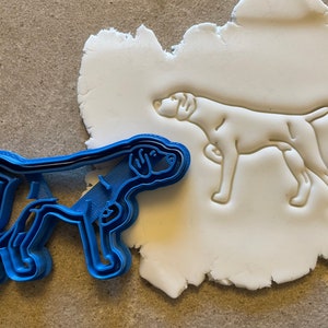 May include: A blue plastic cookie cutter in the shape of a dog, next to a white fondant cookie with an impression of the dog.