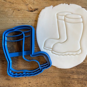 May include: A blue plastic cookie cutter shaped like a pair of rain boots. The cutter is shown next to a white piece of modeling clay that has been pressed with the cutter to create an impression of the boots.