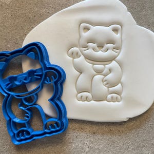 May include: Blue plastic cookie cutter shaped like a cartoon cat with its paw raised in a beckoning gesture. The cutter is next to a white fondant impression of the same cat.