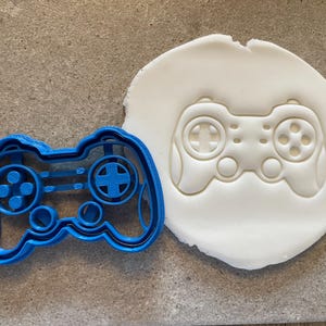 Może przedstawiać: Niebieska plastikowa foremka do ciastek w kształcie kontrolera do gier wideo leży obok białego wycięcia z ciasta tego samego kontrolera. Foremka ma szczegółowe przyciski i pady kierunkowe, idealne do tematycznych smakołyków.