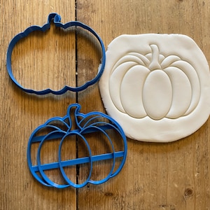May include: Two blue plastic pumpkin cookie cutters and a white pumpkin cookie imprint. The cutters have different designs, one is a simple outline and the other has lines to create a ribbed effect.