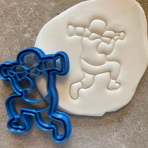 May include: A blue plastic cookie cutter shaped like a person playing a trumpet. The cutter is shown next to a white fondant surface with an impression of the cutter.