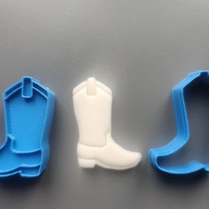 May include: Three cowboy boot-shaped items. A white boot is centered between two blue boot-shaped cookie cutters. The items are arranged on a grey surface. The white boot has a detailed design, while the blue ones are outlines.