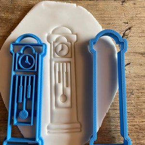 May include: Two blue plastic cookie cutters shaped like a grandfather clock. The cutter on the left is detailed with a clock face, pendulum, and weights. The cutter on the right is a simple outline of the clock.