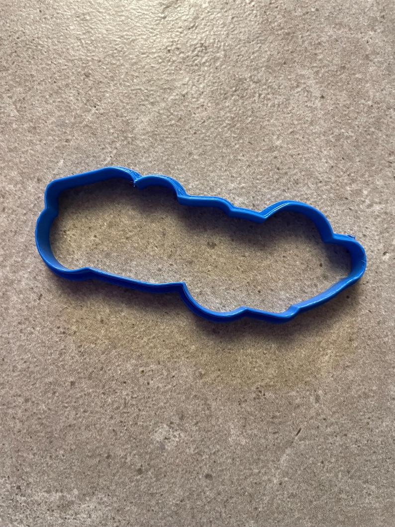 Puede incluir: Un cortador de galletas de pl&aacute;stico azul brillante con forma de nube. El cortador es de una sola pieza con un borde liso y redondeado. Est&aacute; sobre una superficie gris texturizada.