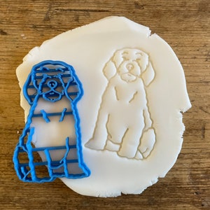Op de afbeelding: Een blauwe plastic koekjesvormer in de vorm van een hond die zit met gekruiste poten. De vormer ligt op een wit fondant oppervlak.