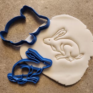 May include: Two blue plastic cookie cutters shaped like rabbits, one with a more detailed design, and a white fondant cookie with a rabbit impression.