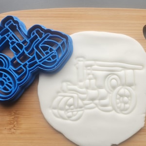 May include: A blue plastic cookie cutter in the shape of a tractor sits next to a white fondant cutout of the same tractor design. The cutter and fondant are on a wooden cutting board. The tractor has large wheels and detailed features.