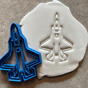 May include: Blue plastic cookie cutter shaped like a fighter jet. The cutter is shown on top of a white fondant surface with an impression of the jet.