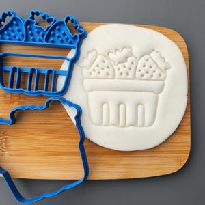May include: A blue plastic cookie cutter and a white fondant impression of strawberries in a basket. The cutter is in the shape of a basket of strawberries. The items are on a wooden chopping board.