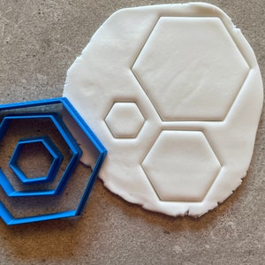 May include: A set of three blue plastic hexagon cookie cutters, with the largest cutter on top of the others. The cutters are next to a piece of white fondant with three hexagon shapes cut out.