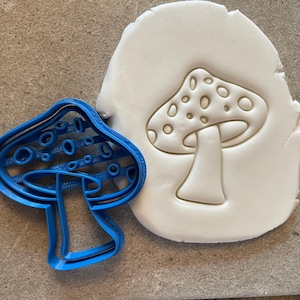 Może przedstawiać: Niebieska plastikowa foremka do ciasteczek w kształcie grzyba z białymi kropkami. Foremka leży na powierzchni białego fondantu z odciskiem grzyba.