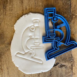 May include: A blue plastic microscope-shaped cookie cutter and a white dough cutout of a microscope. The cutter is a detailed outline of a microscope. The dough shows the impression of the microscope, perfect for baking themed biscuits.